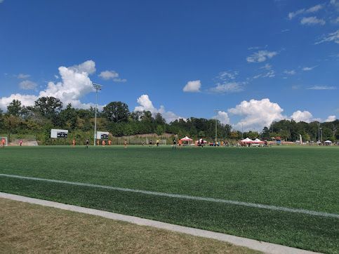 Wide green sports field under a blue sky with clouds, trees, and tents in the distance