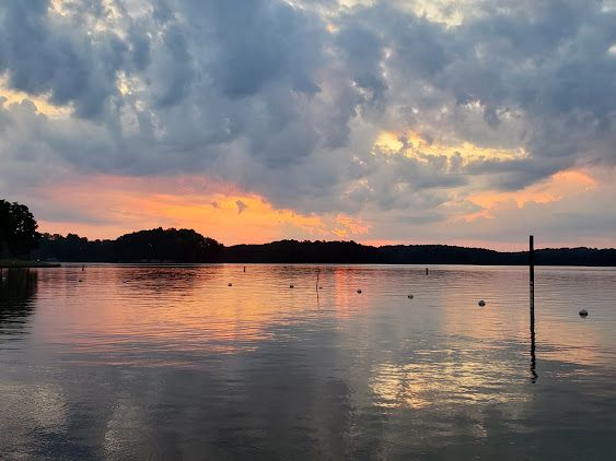 Calm lake at sunset with pink-orange clouds reflecting on the water and dark tree-lined shore