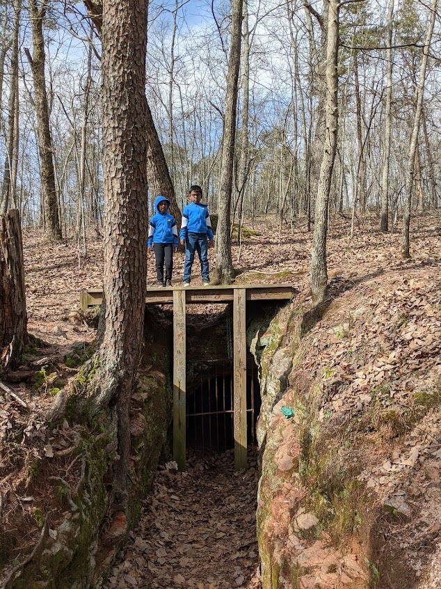 Two hikers in blue jackets stand by a wooden bridge over a narrow rocky ravine in a leafless forest.