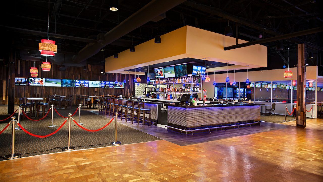 Empty casino floor with a brightly lit bar and slot machines behind red rope barriers.