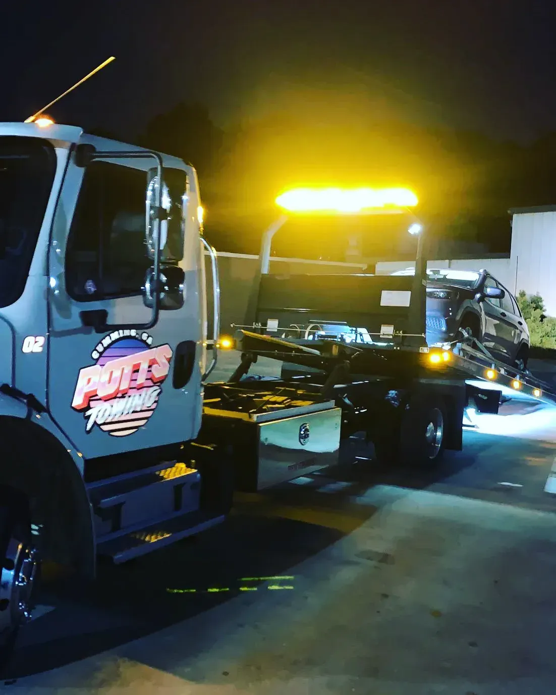 Tow truck at night with bright yellow lights, hauling a damaged car on the road
