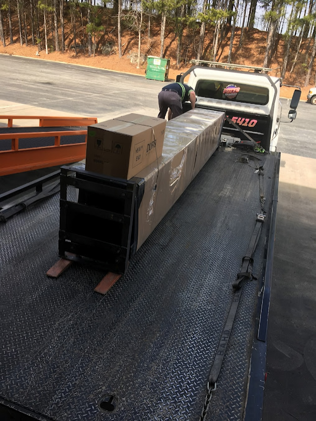 Worker loading a long wrapped package onto a flatbed truck on a roadside bridge.