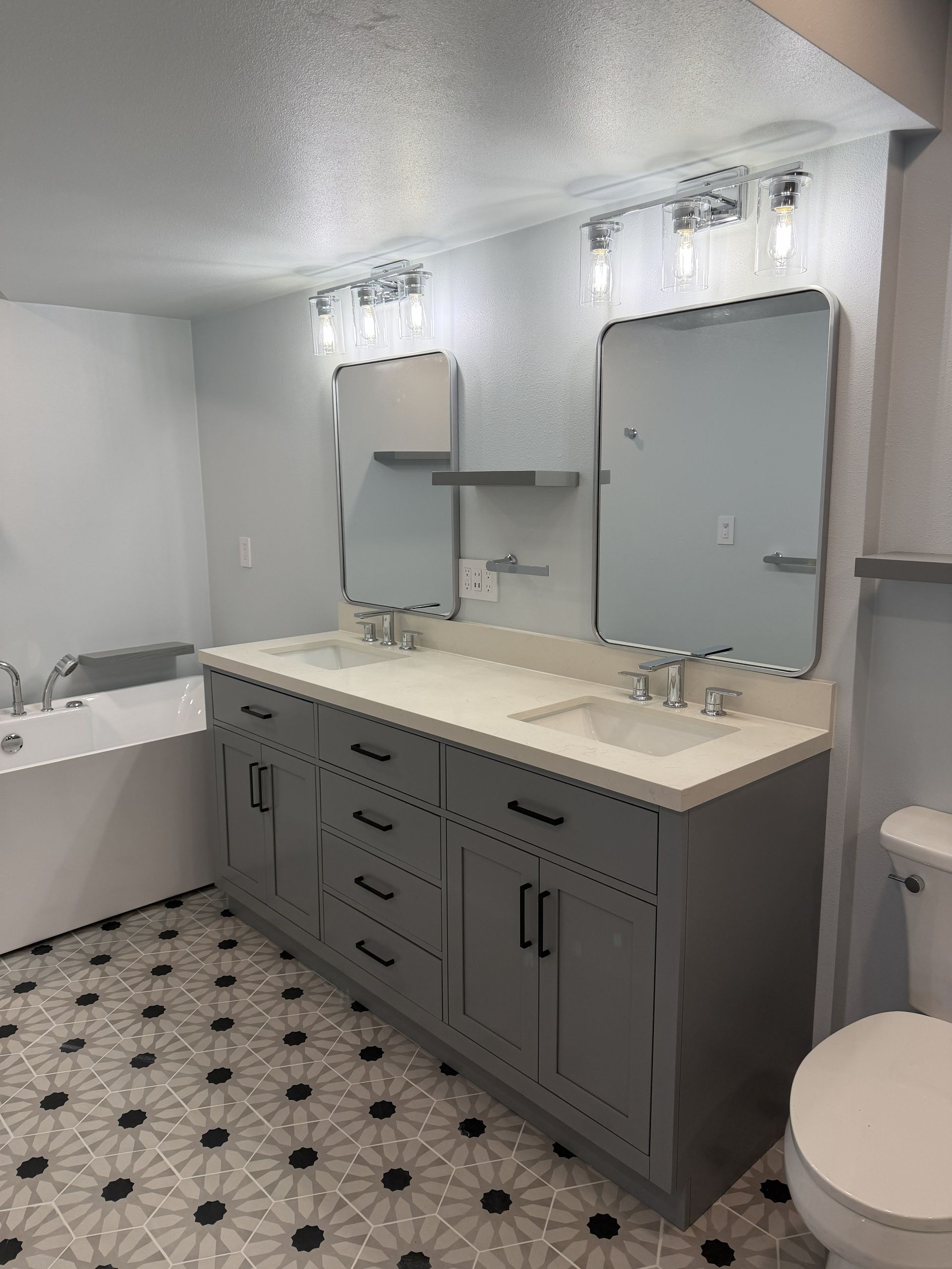 Modern bathroom with gray vanity, double mirrors, white tub, and black-and-white tile floor