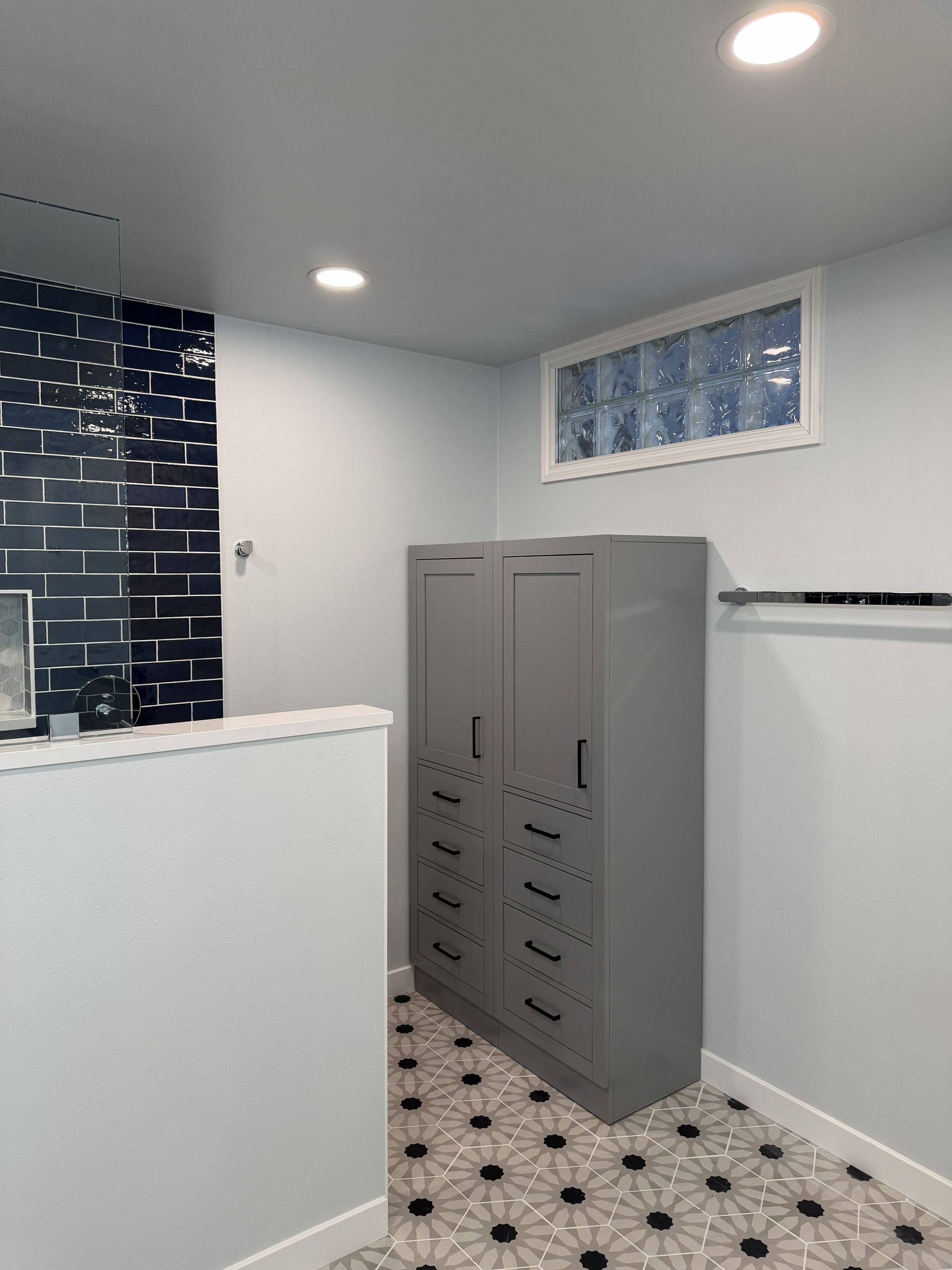 Small bathroom with gray cabinets, white walls, patterned tile floor, and a window.