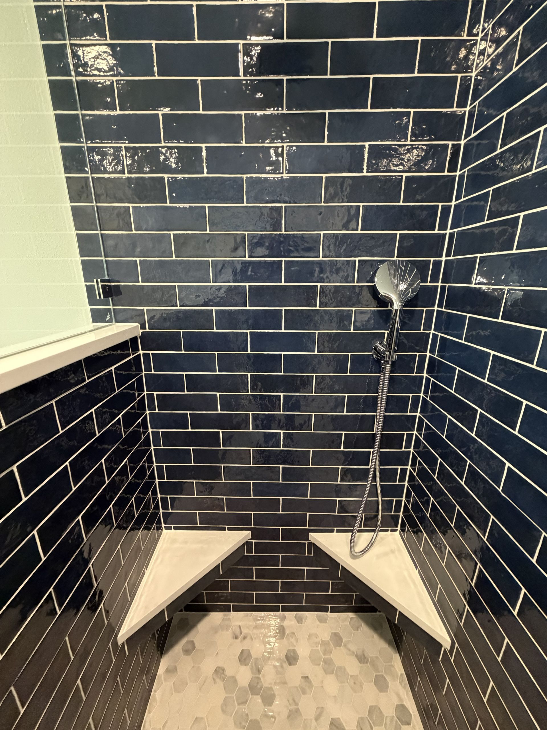 Empty shower stall with dark blue tile, white corner shelves, and a showerhead on the right wall.