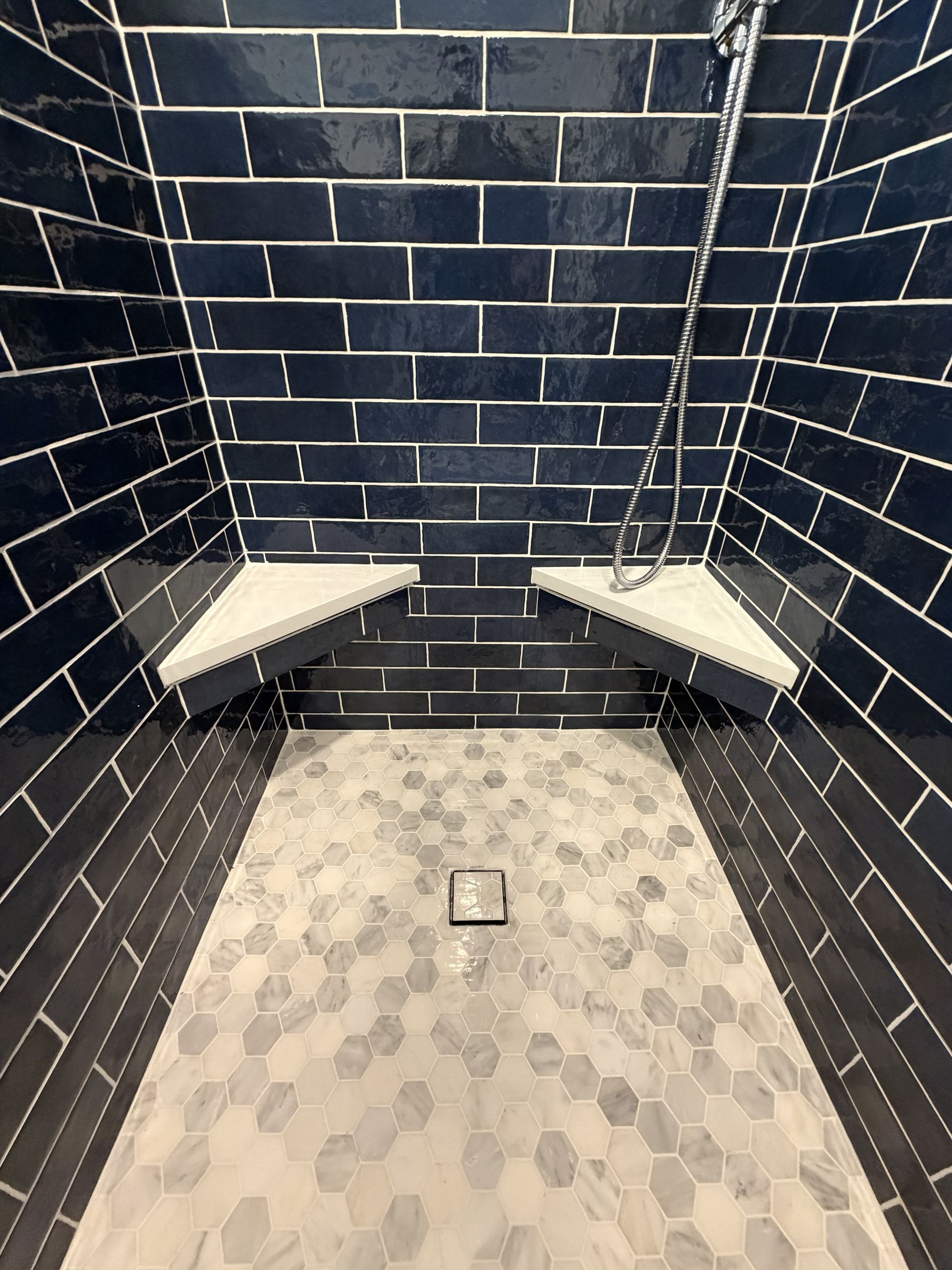 Accessible shower stall with dark blue tile walls, built-in corner benches, and a small floor drain.