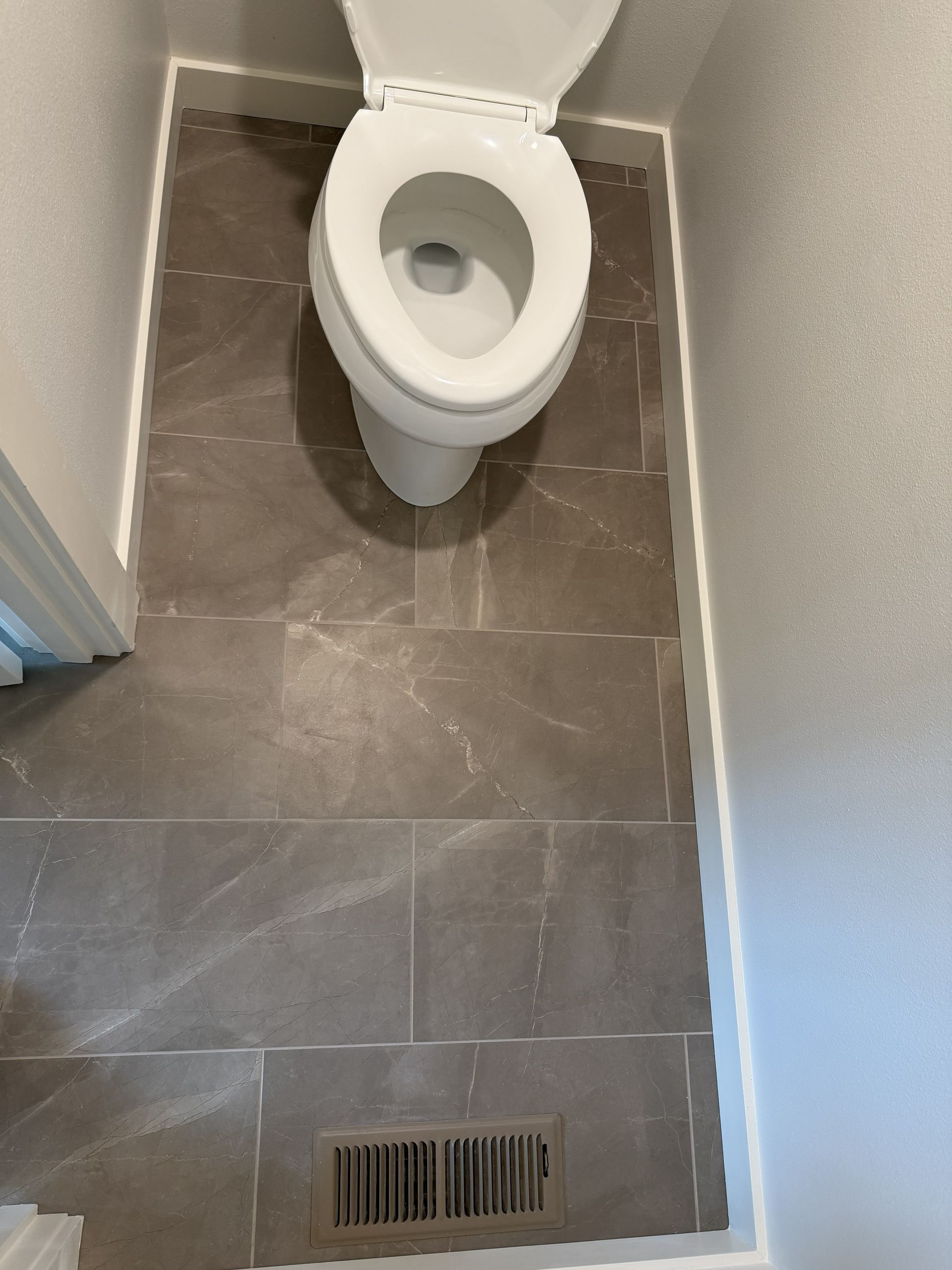 Small bathroom with a white toilet over gray tile flooring and a floor vent.