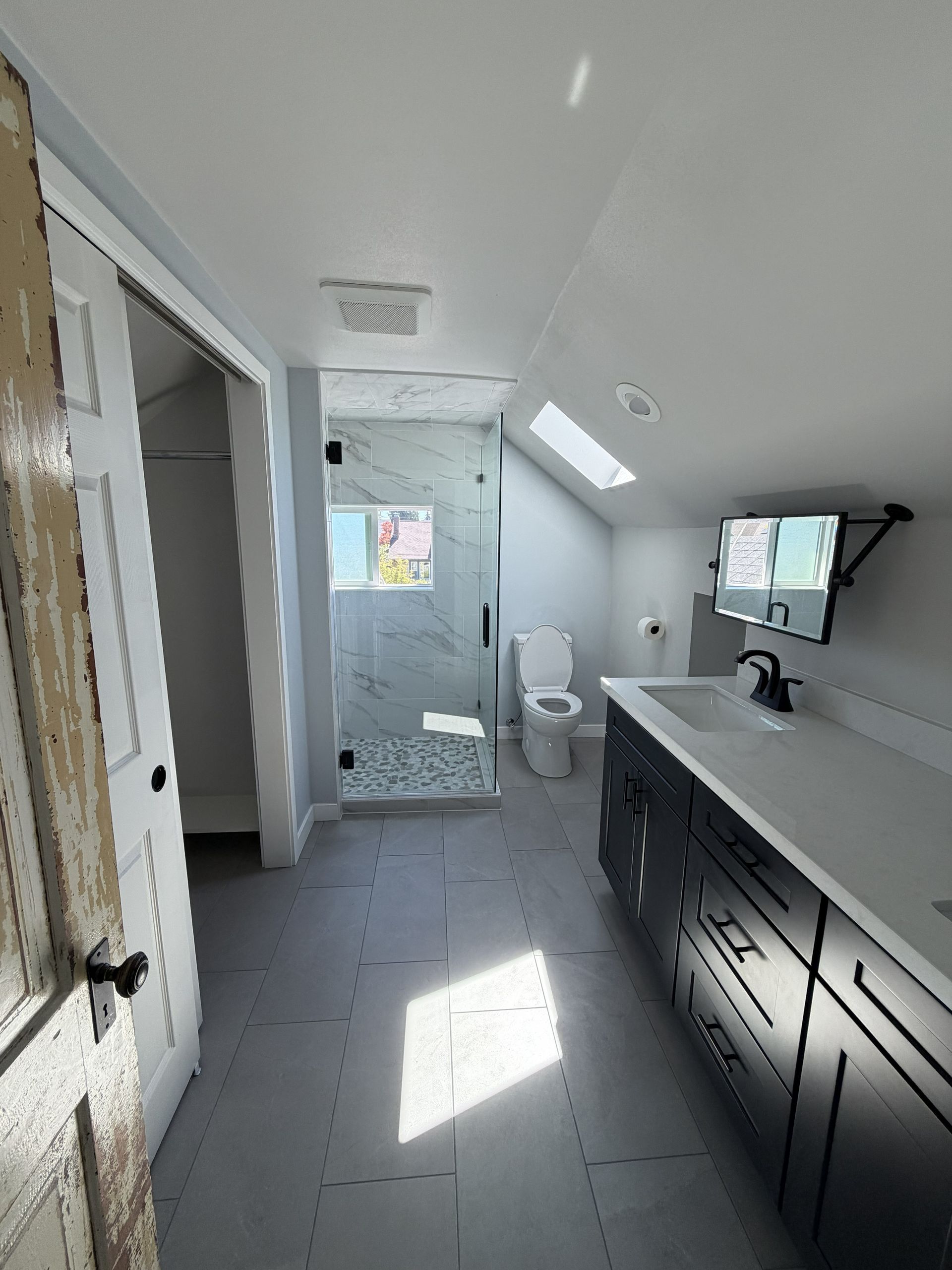 Modern bathroom with gray tile, shower, toilet, and dark vanity under a slanted ceiling.