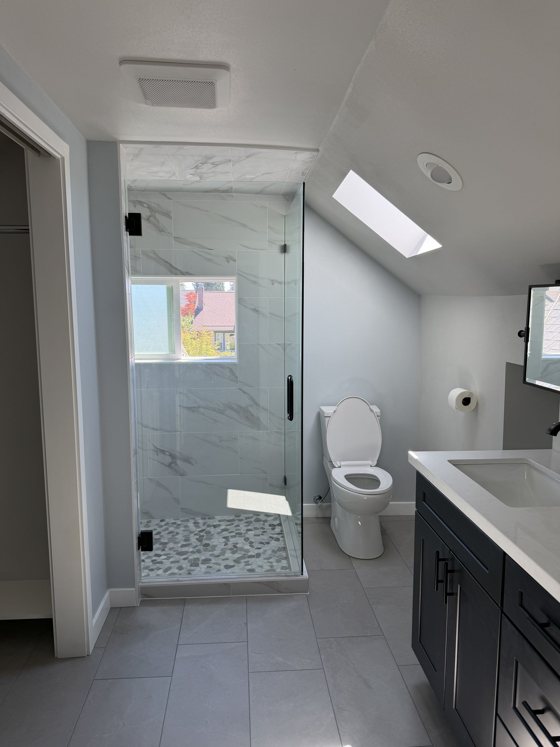Modern bathroom with glass shower, toilet, skylight, and dark vanity under a sloped ceiling