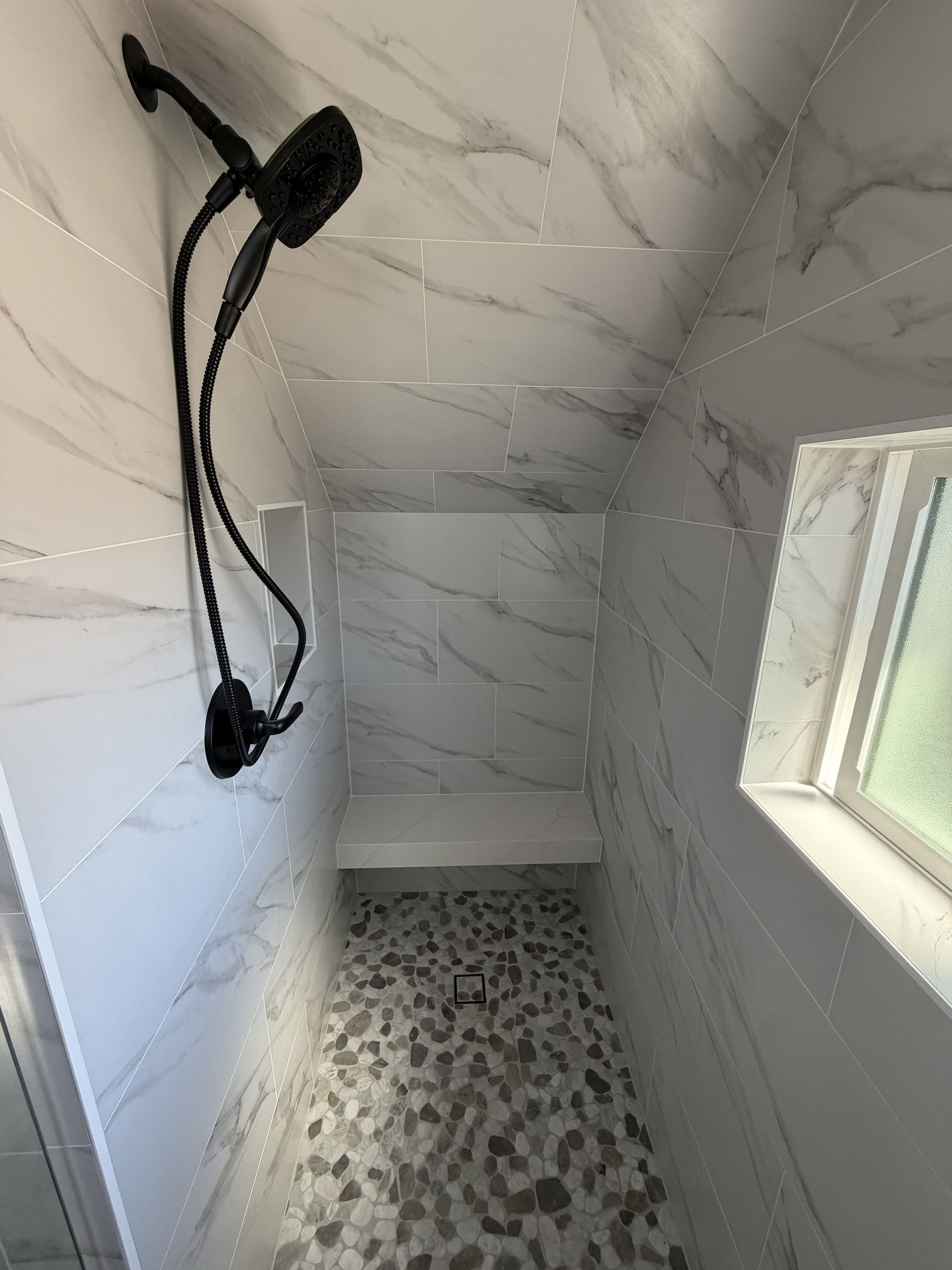 Tiled shower stall with two mounted black showerheads and a small frosted window.