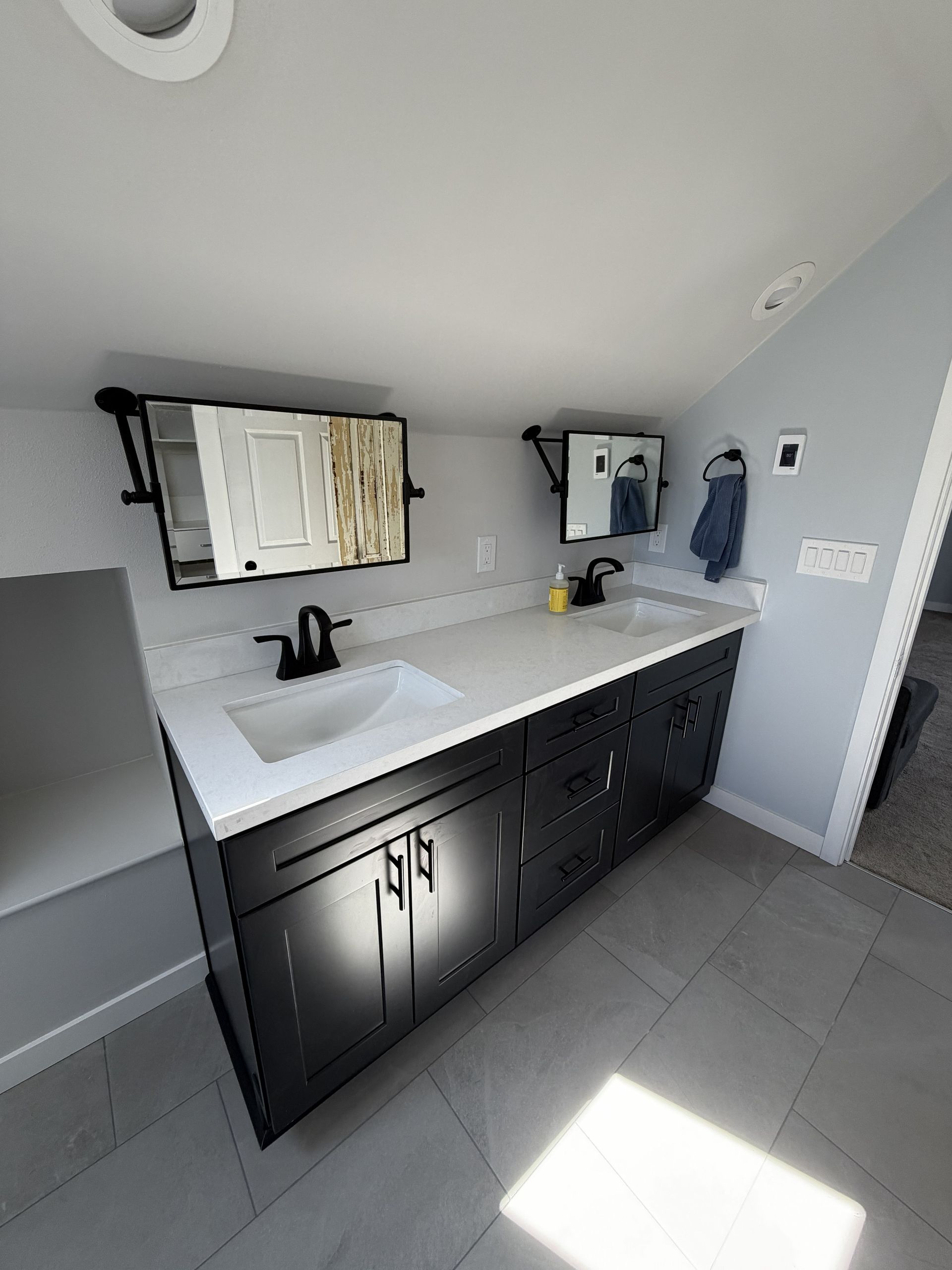 Modern bathroom vanity with double sinks, dark cabinets, black mirrors, and sunlight on gray tile floor