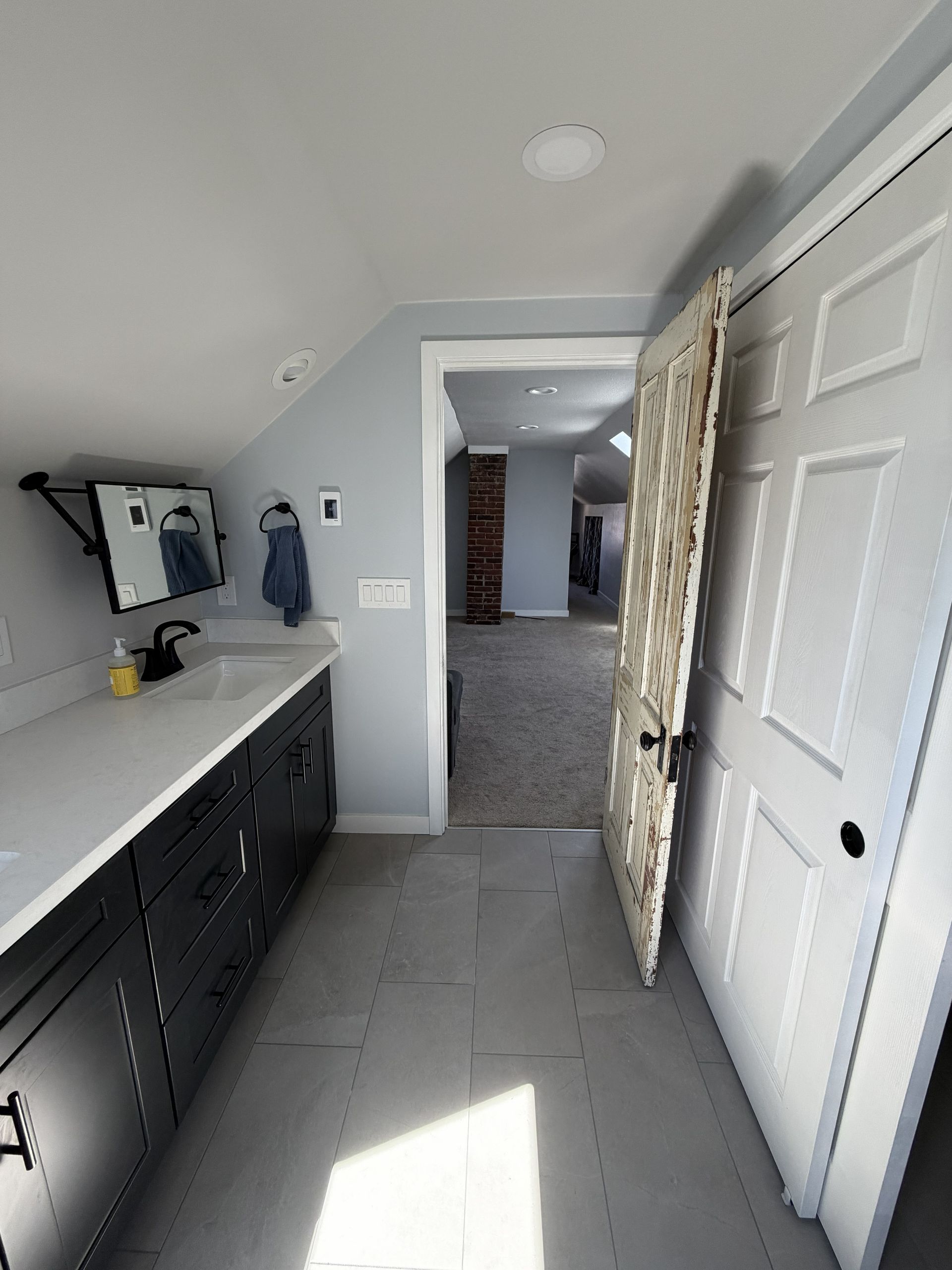 Narrow hallway with white and black bathroom vanity, open door, and bright sunlight on tiled floor