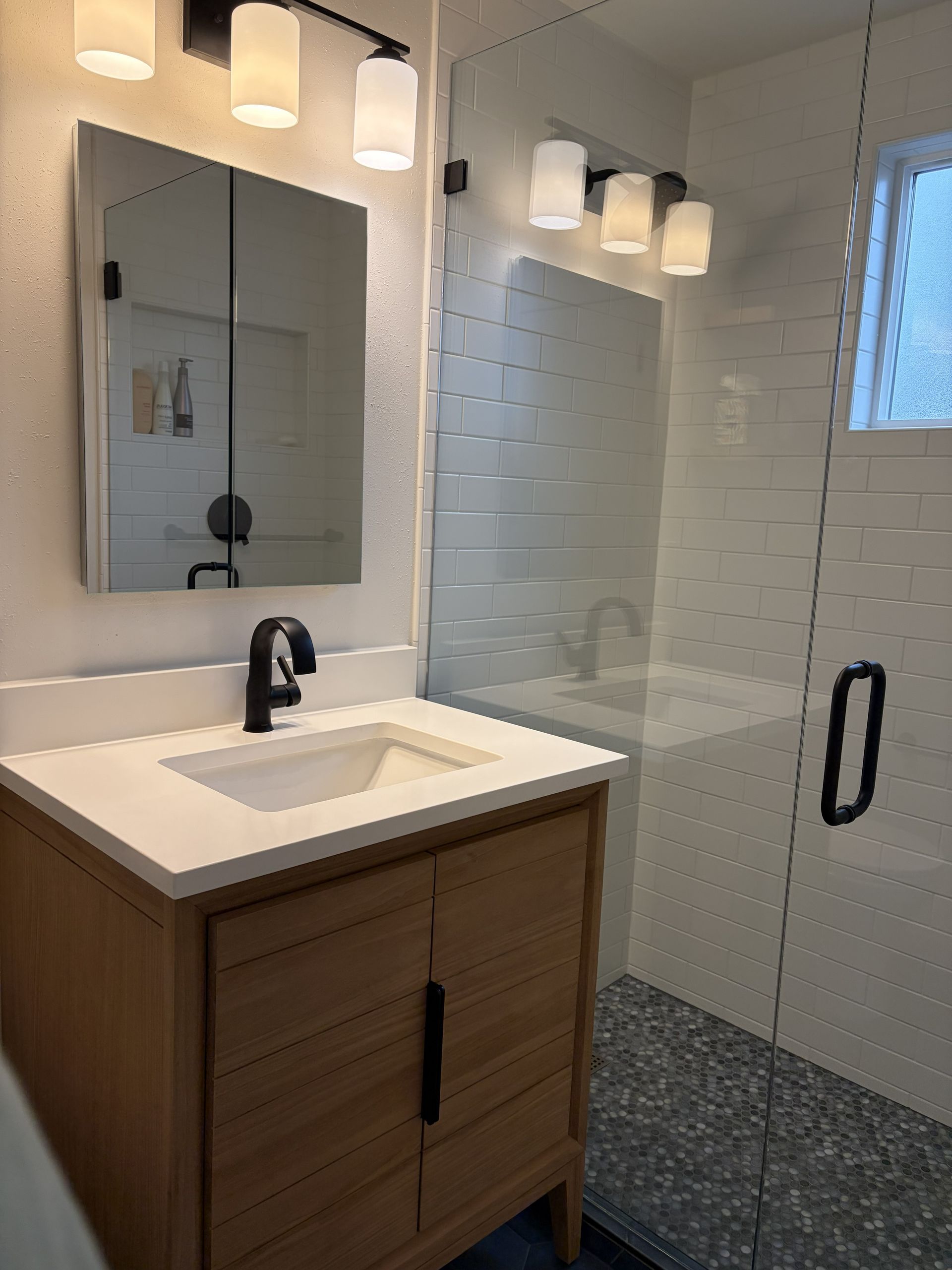 Modern bathroom with wood vanity, white sink, mirror, sconces, and tiled shower behind glass door