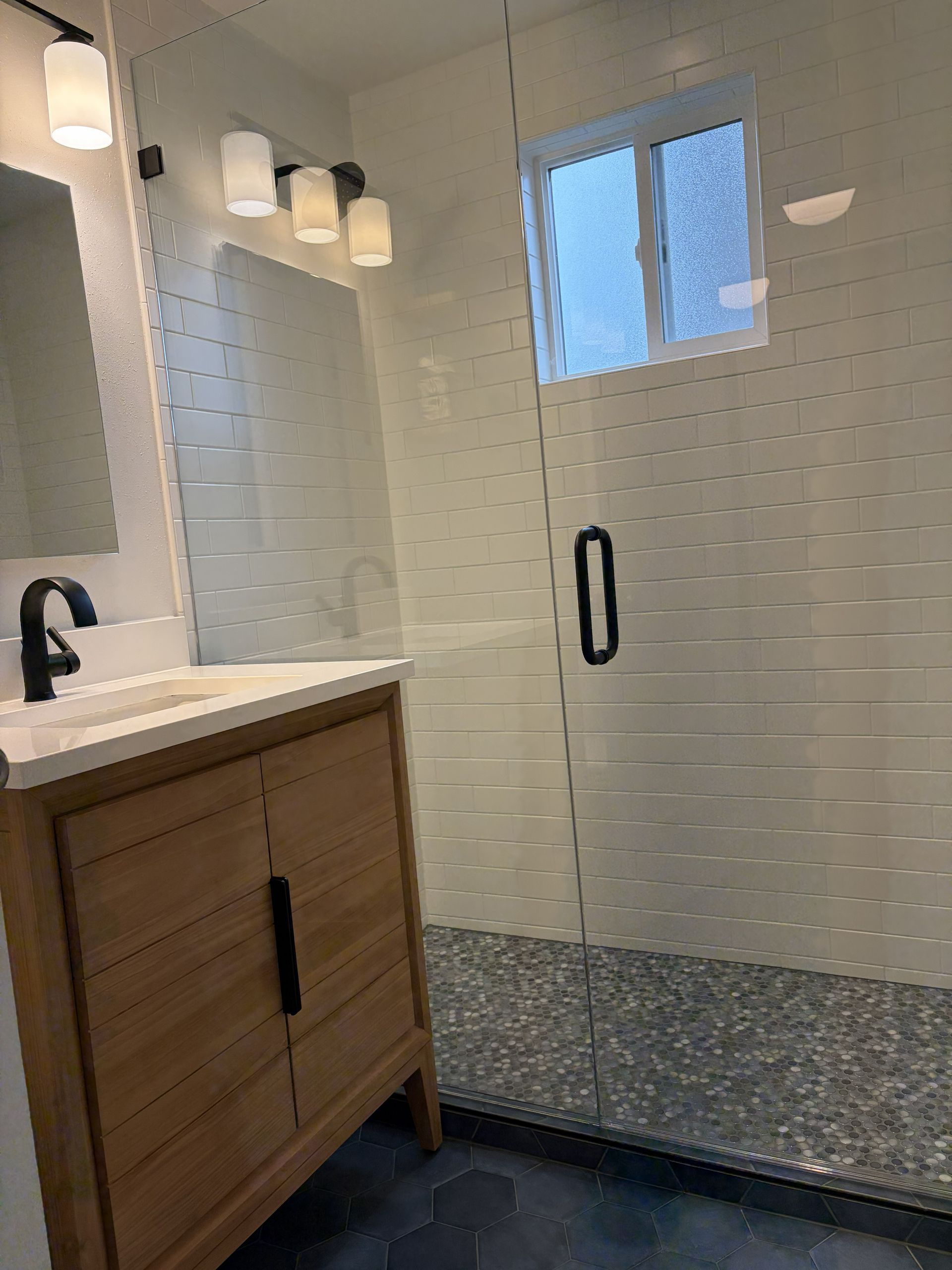 Modern bathroom with wood vanity, black fixtures, white tiled shower, and a frosted window