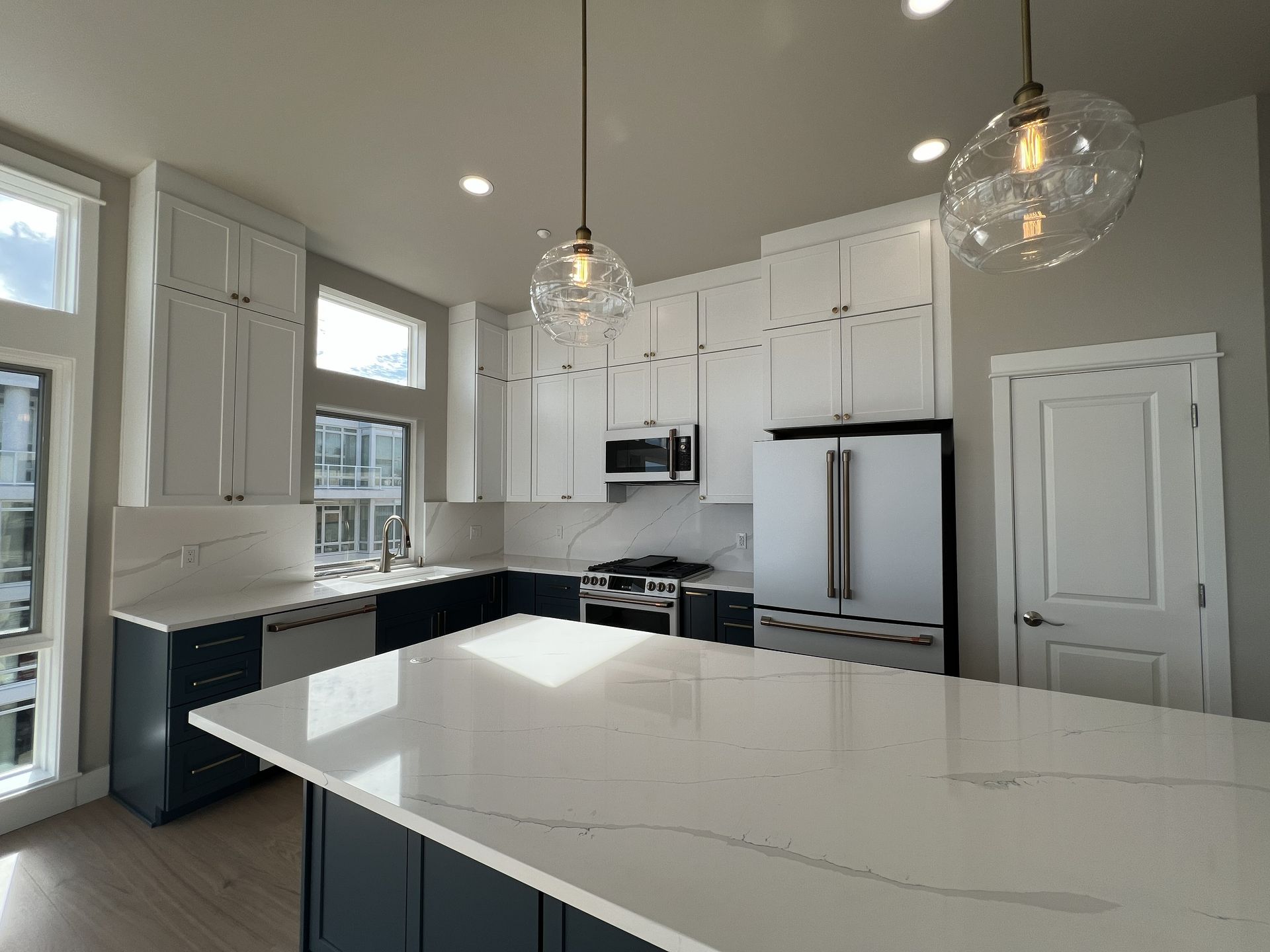 Modern white kitchen with large island, pendant lights, stainless appliances, and tall windows