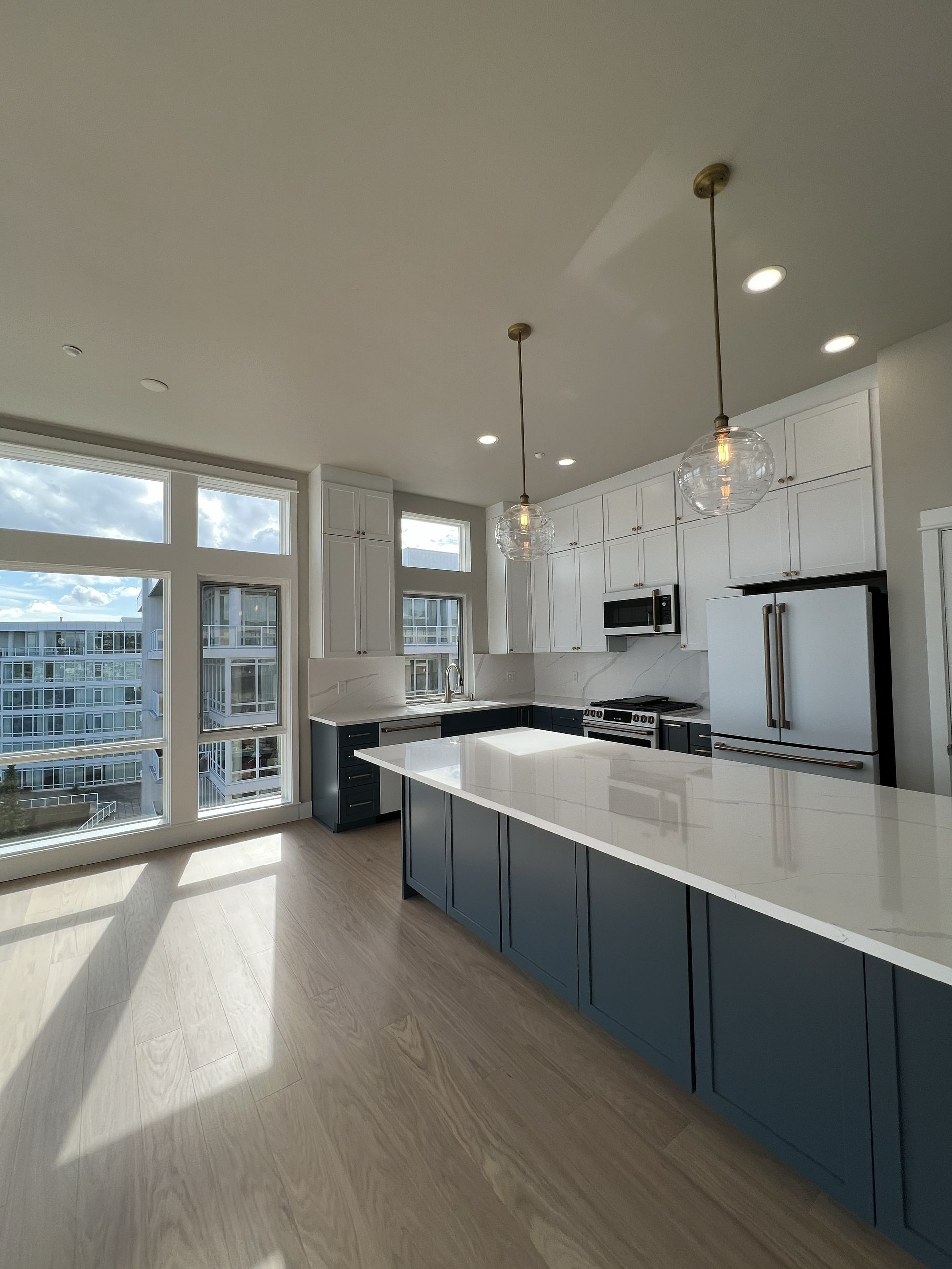 Bright modern kitchen with large island, white cabinets, blue lower cabinets, and tall windows.