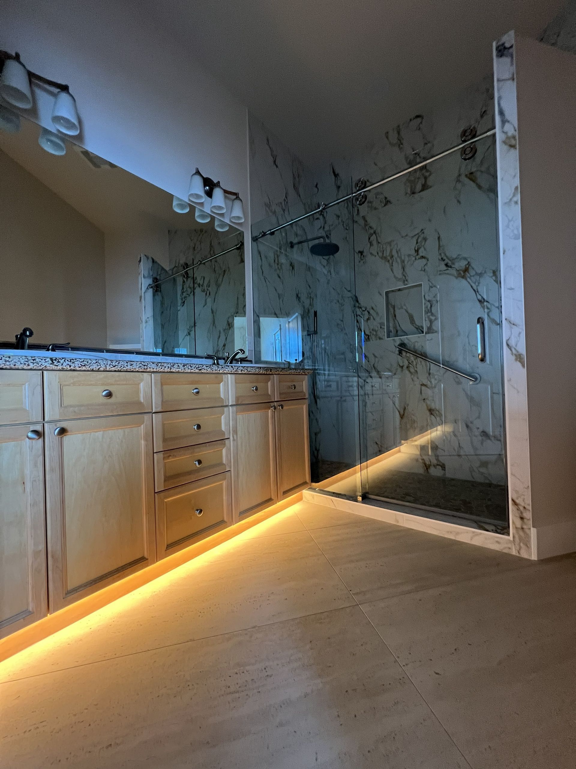 Modern bathroom with wood vanity, large mirror, and glass shower with lit floor strip lighting.