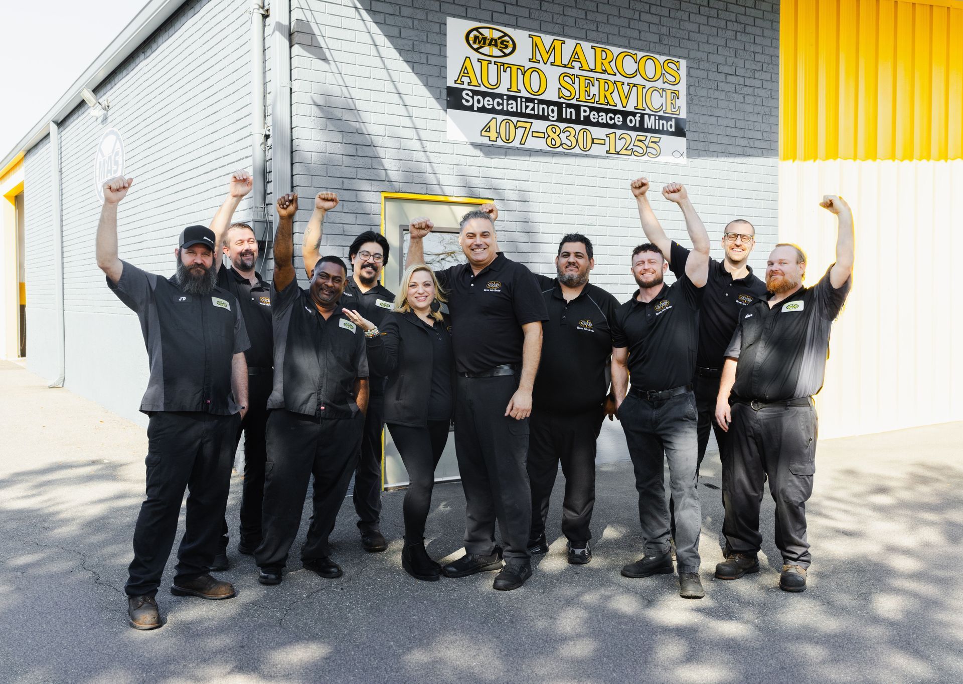 Group of auto shop employees celebrating in front of their building, arms raised. | Marcos Auto Service