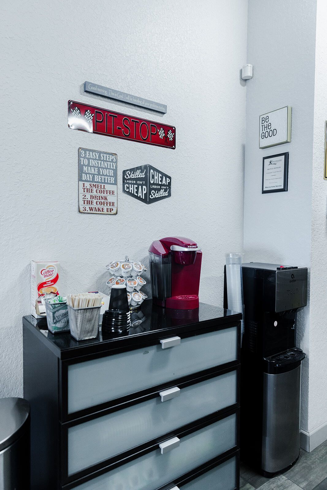 Coffee station: red Keurig, water dispenser, black cabinet with silver handles, decorative wall art. | Marcos Auto Service