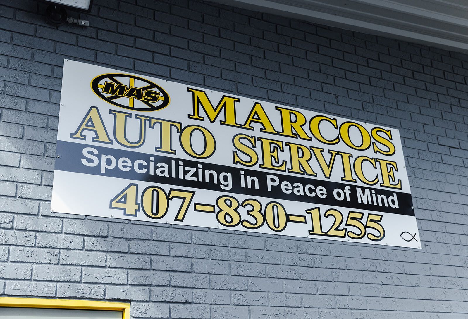 Sign for Marcos Auto Service, with yellow and black text on a white background, hanging on a gray brick wall; phone number 407-830-1255. | Marcos Auto Service