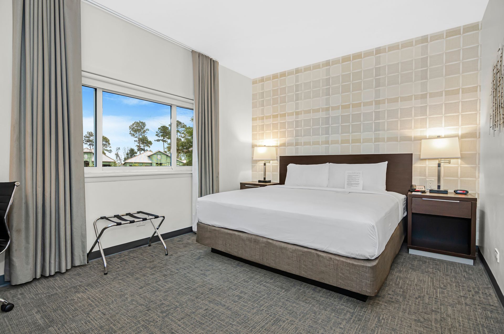 A hotel room with a king-sized bed, grey curtains, bedside tables with lamps, and a luggage rack near a large window.