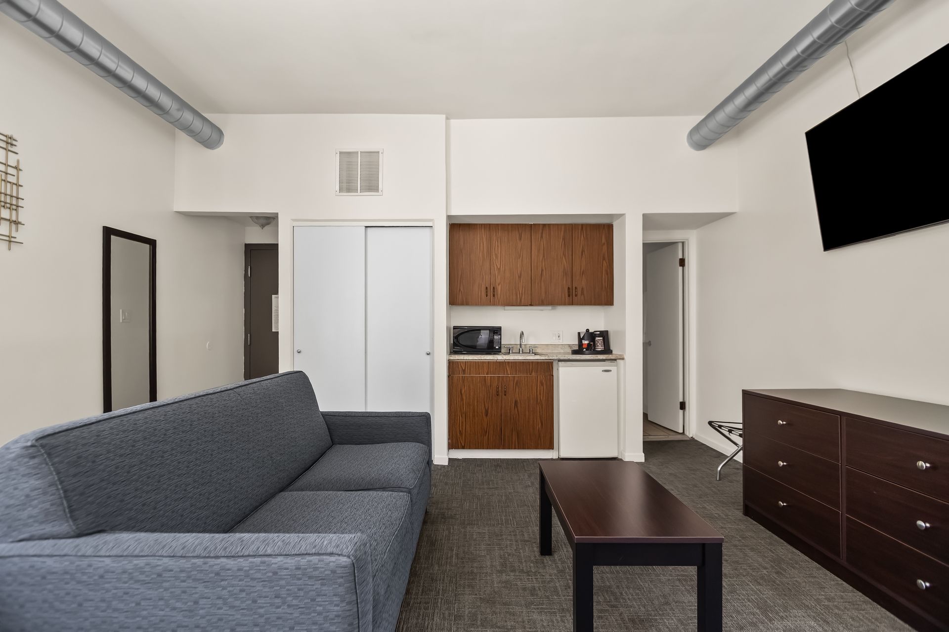 Small furnished living room with gray sofa, coffee table, dresser, and kitchenette.