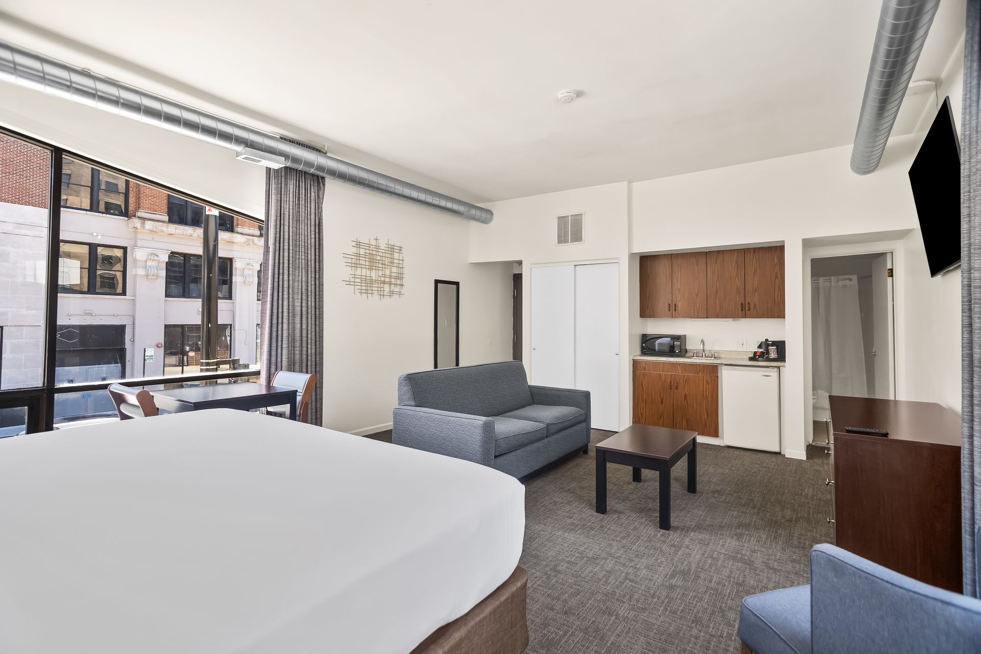 Modern hotel room with a large bed, sofa, desk, and window view of nearby buildings