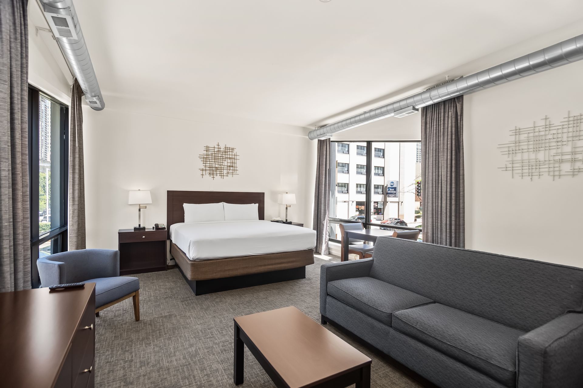 Modern hotel room with a king bed, gray sofa, desk, and balcony doors overlooking city buildings. Pull Out Sofa
