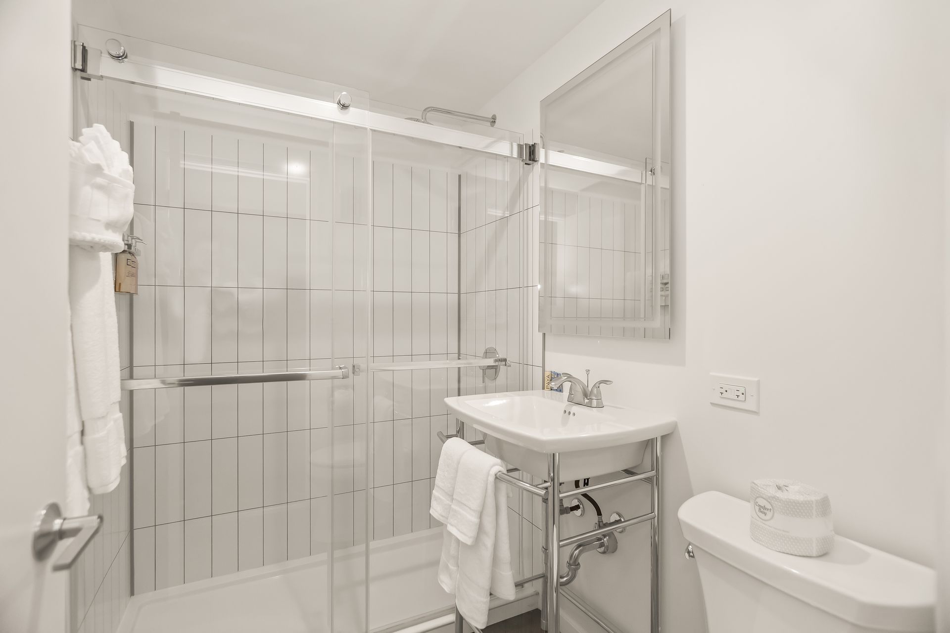 Bright white bathroom with glass shower, pedestal sink, toilet, and large mirror.
