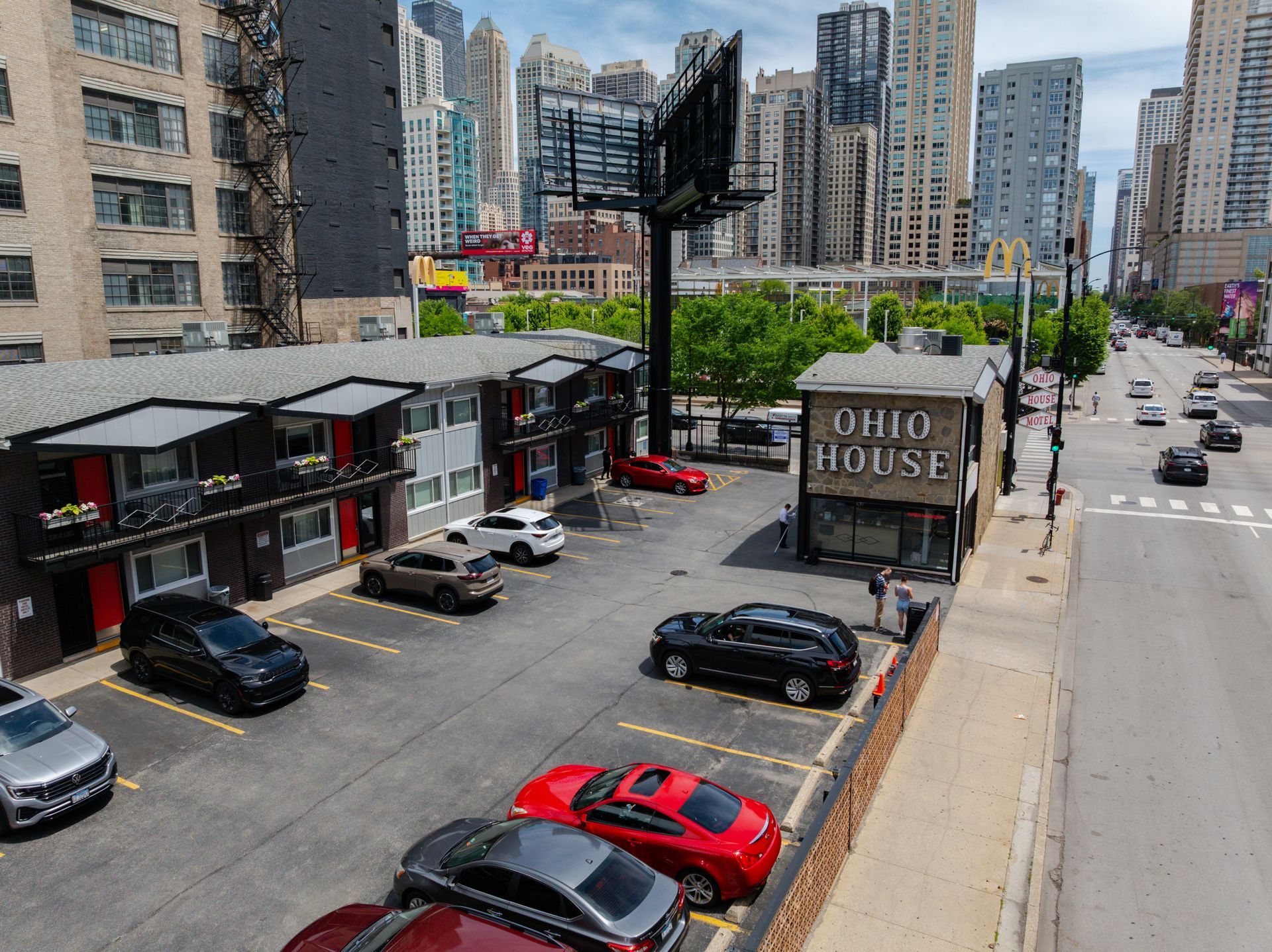 Urban street with parked cars beside low shops and tall apartment buildings on a sunny day