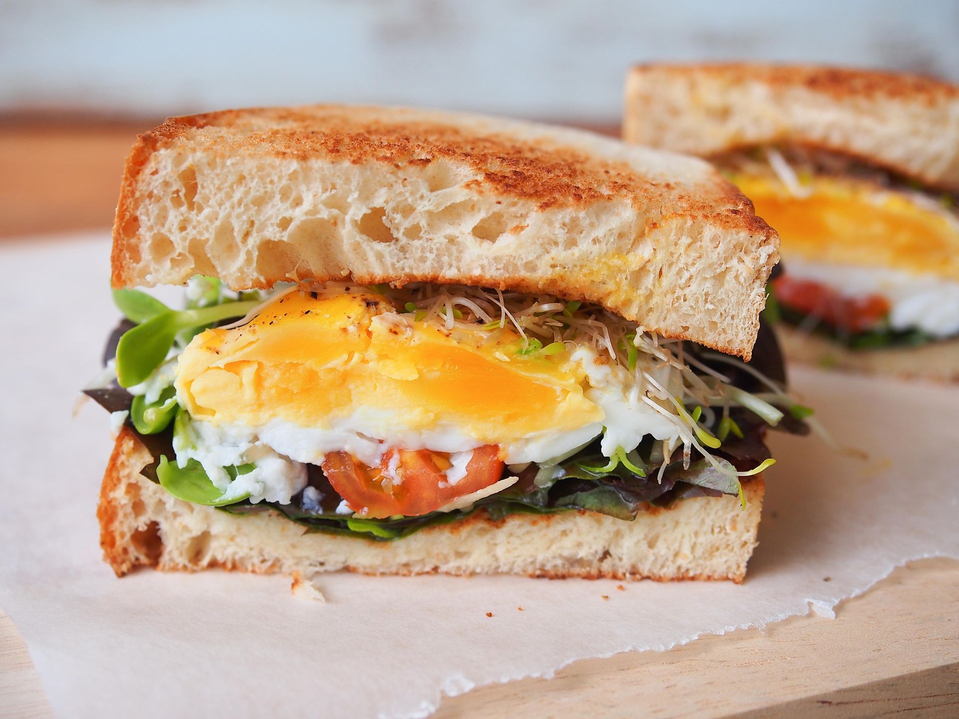A toasted sandwich with a fried egg, sprouts, and sliced tomatoes, sitting on parchment paper.