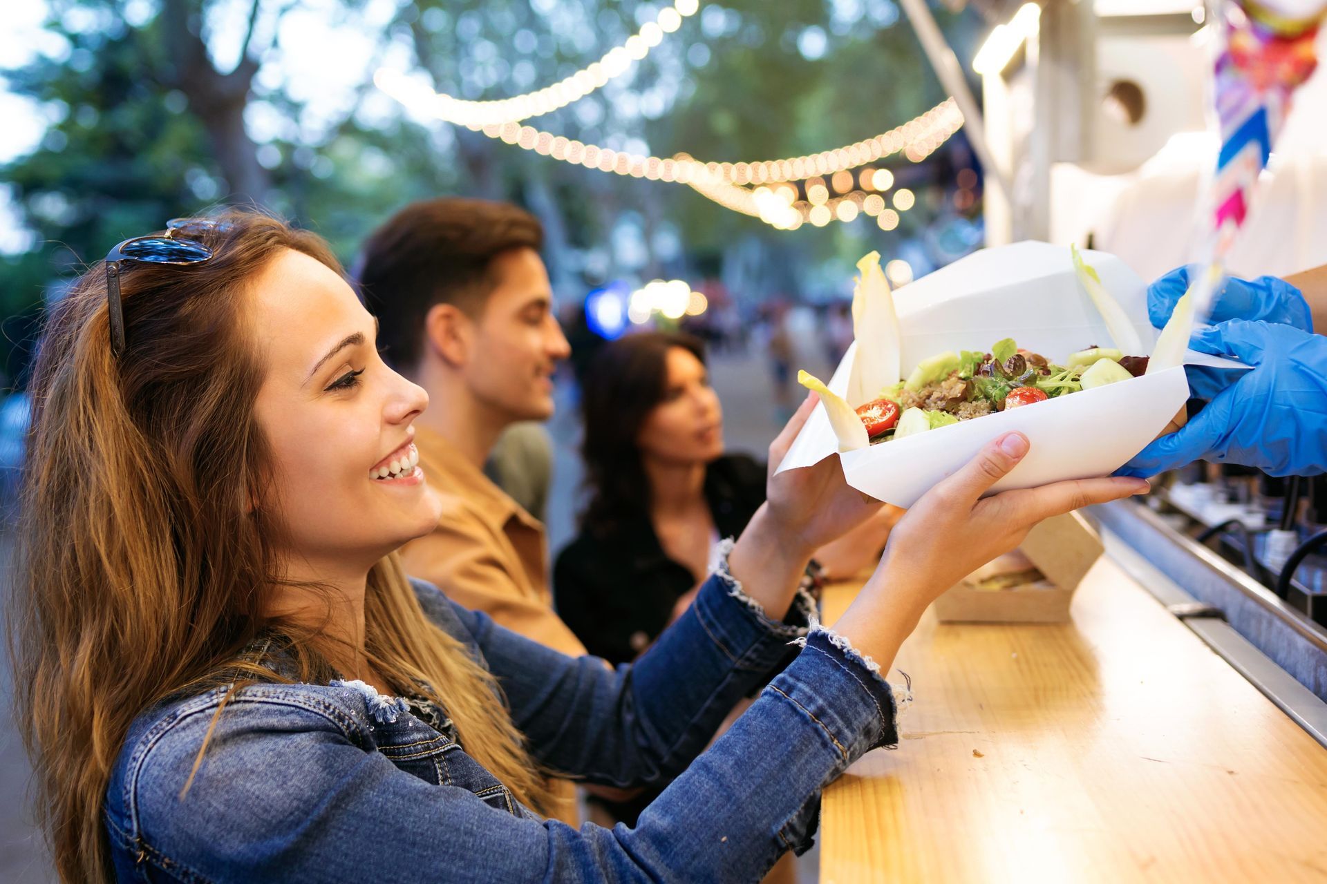 Woman receiving food from a food truck, smiling. Others wait in line. Outdoors.