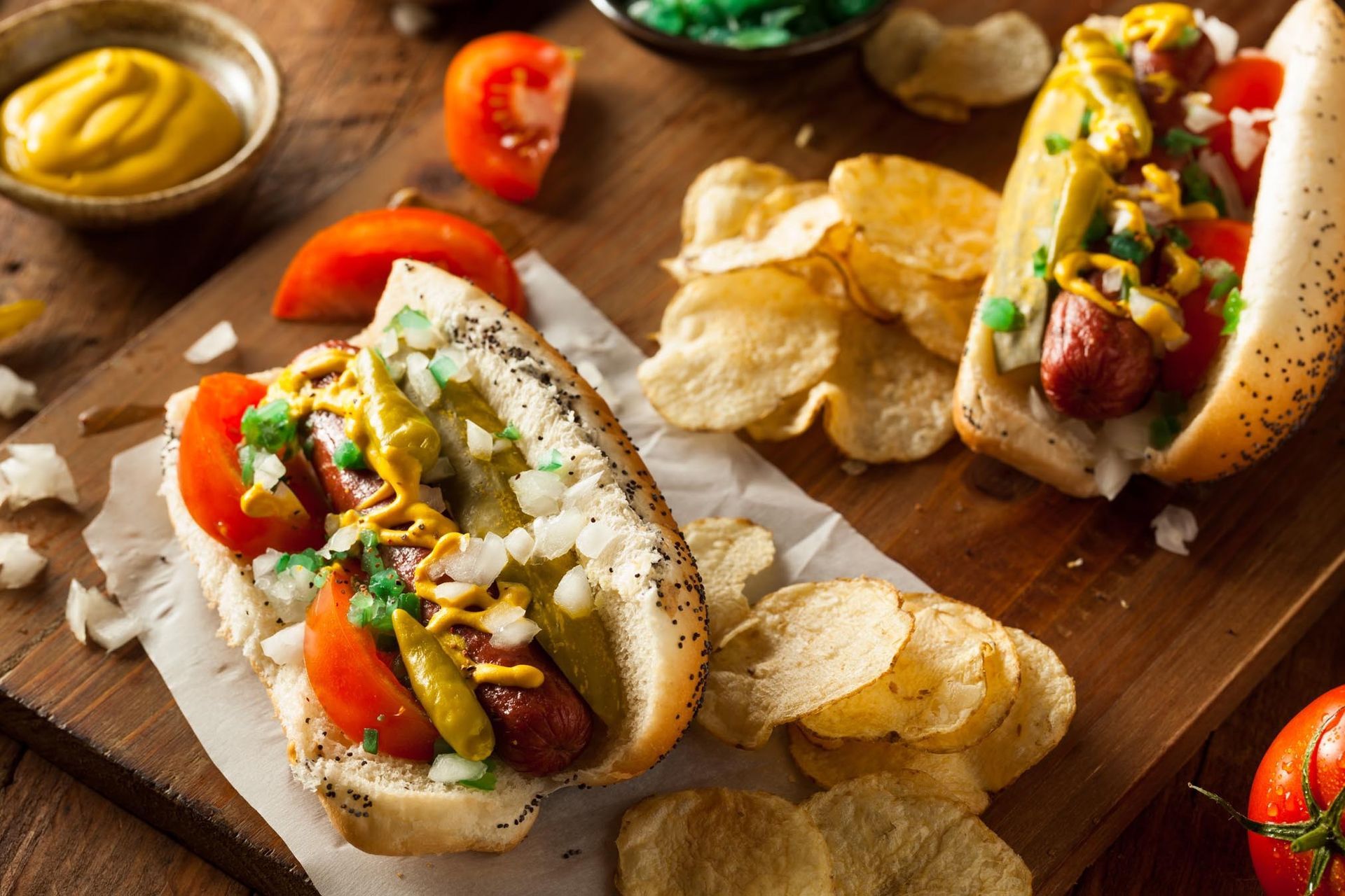 Hot dogs in poppy seed buns with toppings, chips, tomatoes, and mustard on a wooden board.
