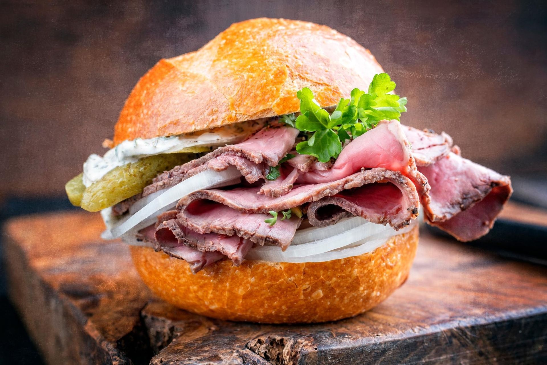 A roast beef sandwich with onion, pickles, and herbs on a crusty bun, served on a rustic wooden board.