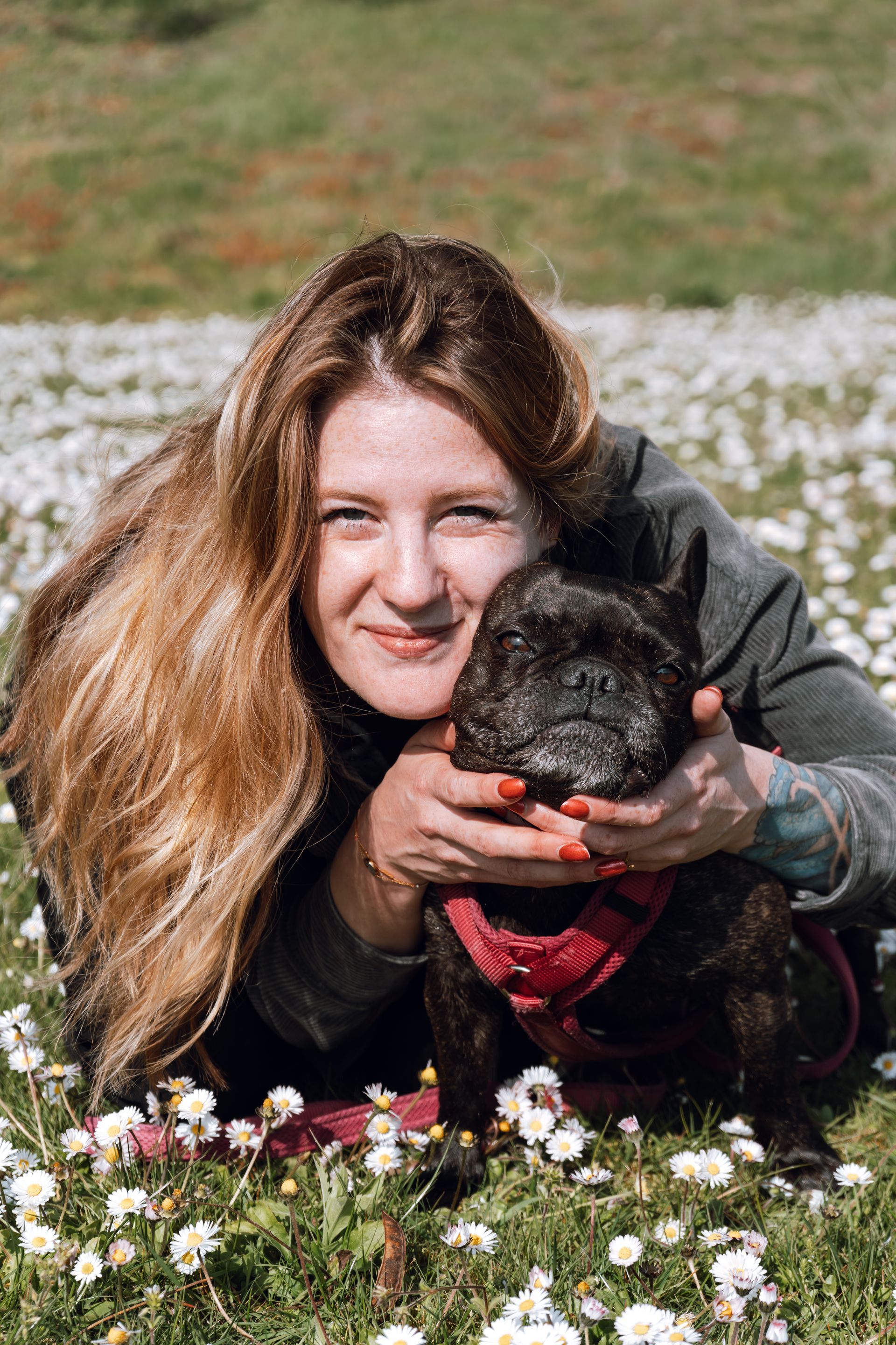 Woman lying in grass, smiling and holding a small black dog with a red bandana.