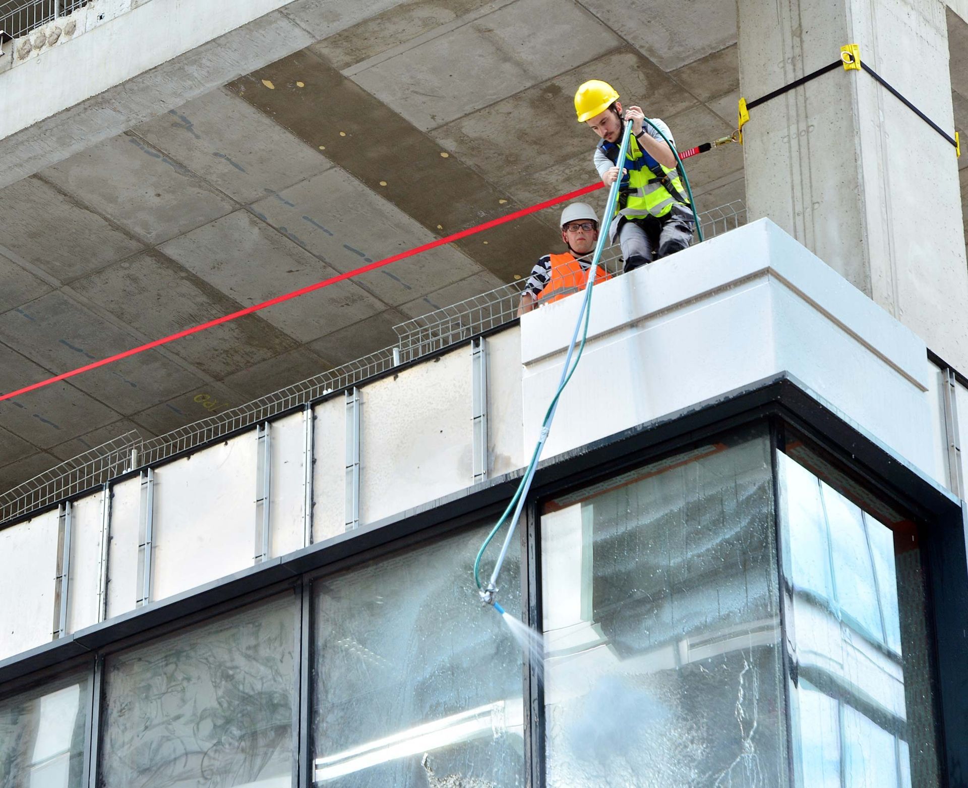 Workers on a building ledge washing windows with a spray nozzle. One wears a harness and helmet.