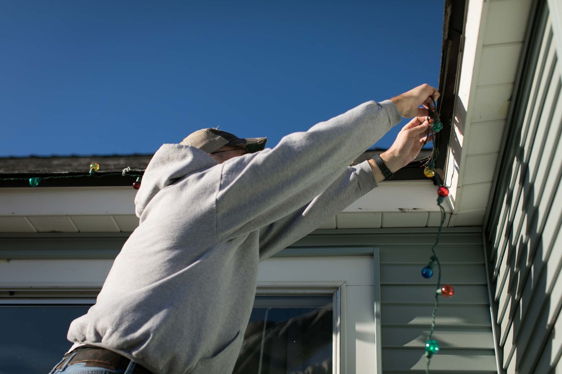 Person on ladder hanging Christmas lights from a house's eaves. Blue sky background.