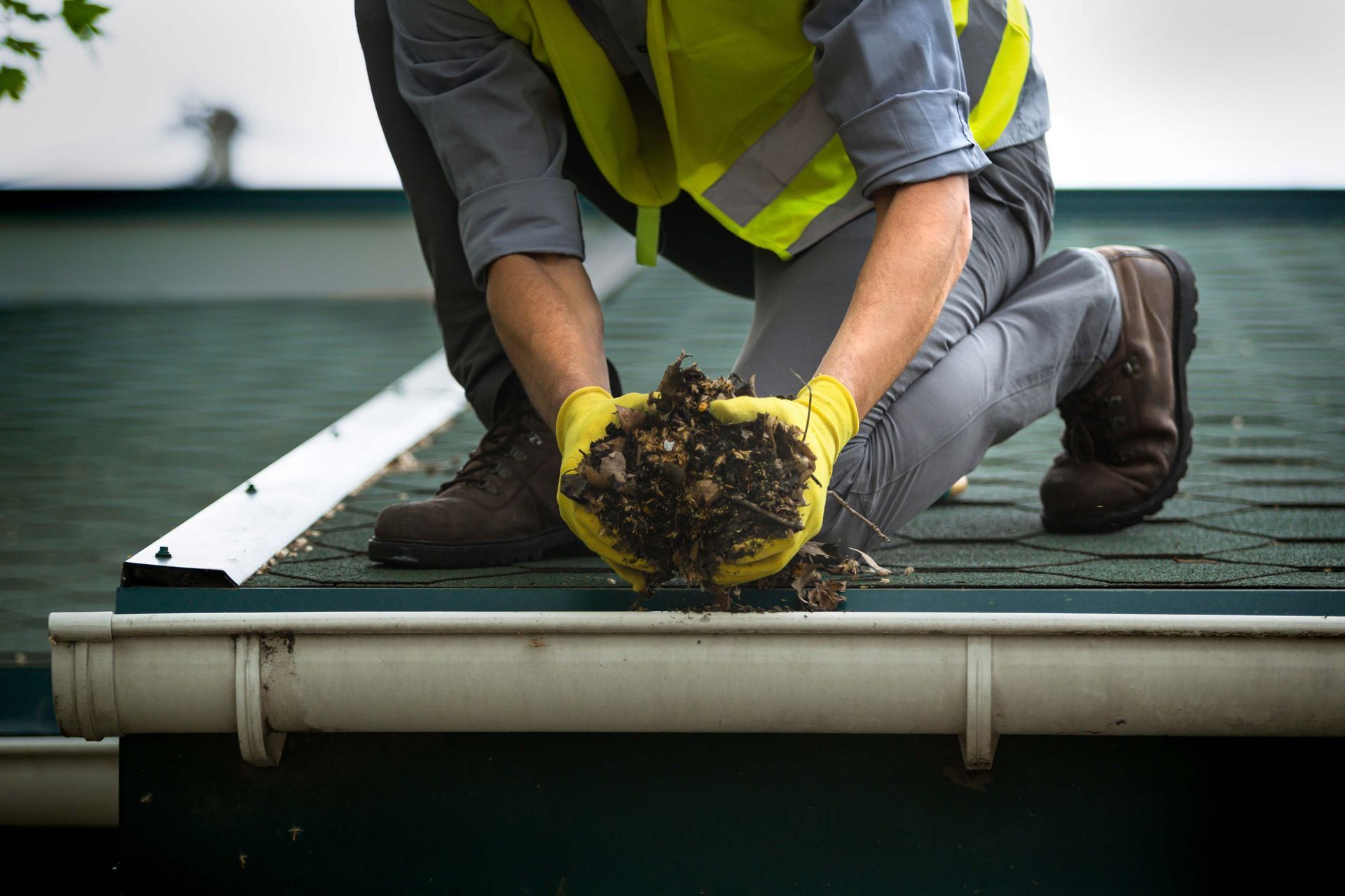 Person cleaning a gutter, holding debris in yellow gloves, on a roof.