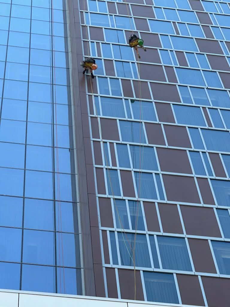 Two window washers on ropes cleaning a tall glass and brown panel building.