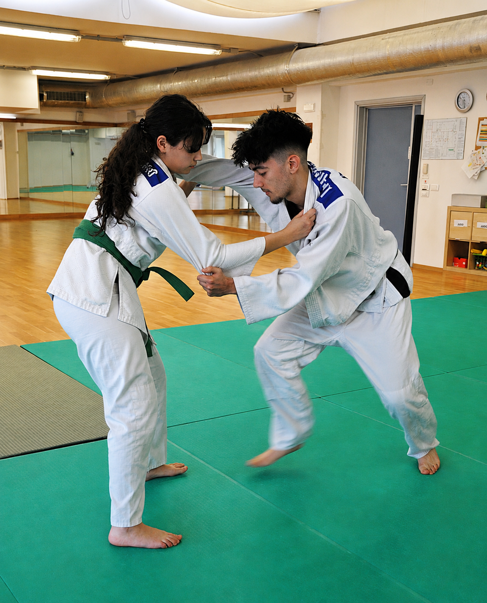 Due persone in uniforme bianca da arti marziali si affrontano in una lotta su un tappeto verde in un dojo.