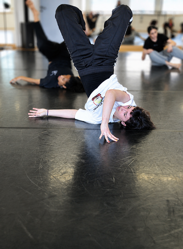 Una persona esegue una posa di breakdance statica sul pavimento di uno studio, con i compagni di classe sfocati sullo sfondo.