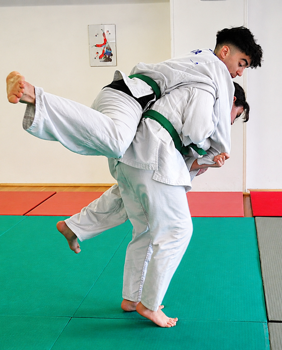 Due atleti di judo con il kimono bianco si esercitano in una proiezione su tappeti verdi e rossi.