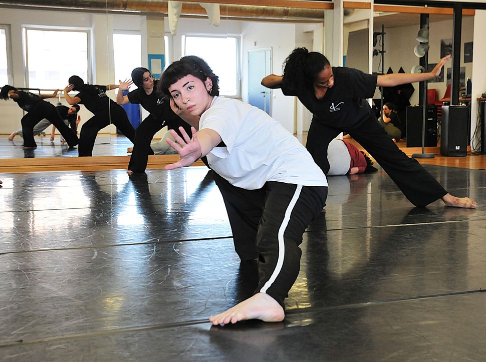 Persone che provano una posa di danza in uno studio, con una persona vestita di bianco al centro in primo piano.