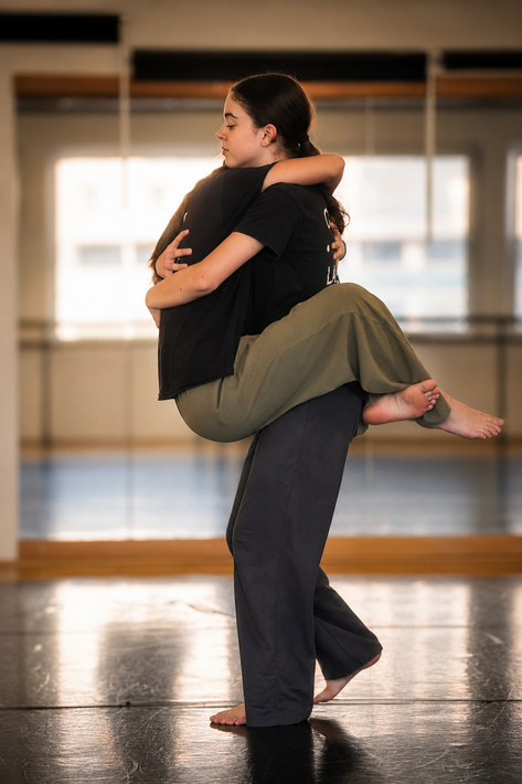 Due ballerine si esercitano in una presa in uno studio con specchi, una vestita di nero, l'altra con pantaloni color oliva.