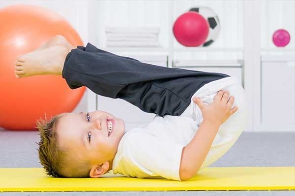 Un bambino è sdraiato su un tappetino giallo, sorridente con una gamba sollevata, in una luminosa stanza dei giochi con delle palle da ginnastica.