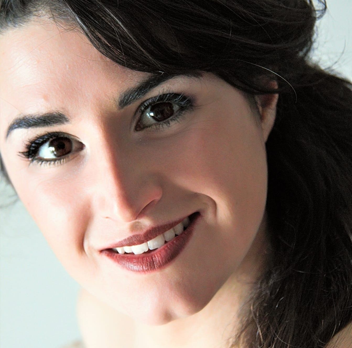 Ritratto ravvicinato di una donna sorridente con capelli scuri e trucco su sfondo chiaro.