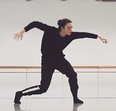Una persona vestita di nero balla in uno studio, sporgendosi in avanti con le braccia tese.