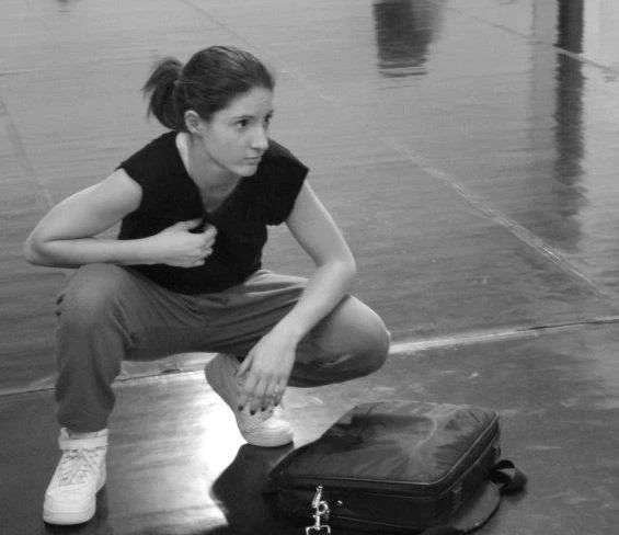 Donna accovacciata sul pavimento di una palestra accanto a una borsa, con lo sguardo rivolto di lato, indossa una maglietta e pantaloni neri.