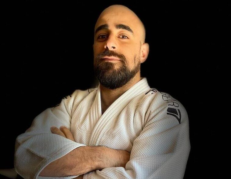 Uomo calvo sorridente con la barba, che indossa un kimono bianco da arti marziali, con le braccia incrociate su sfondo nero.
