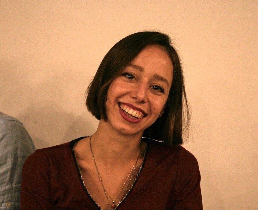 Persona sorridente con capelli corti e scuri, che indossa una maglietta color bordeaux, davanti a una parete beige.